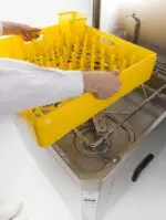 person loading an empty yellow dish rack into the front of a pass through style dishwasher.