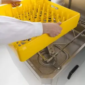 person loading an empty yellow dish rack into the front of a pass through style dishwasher.